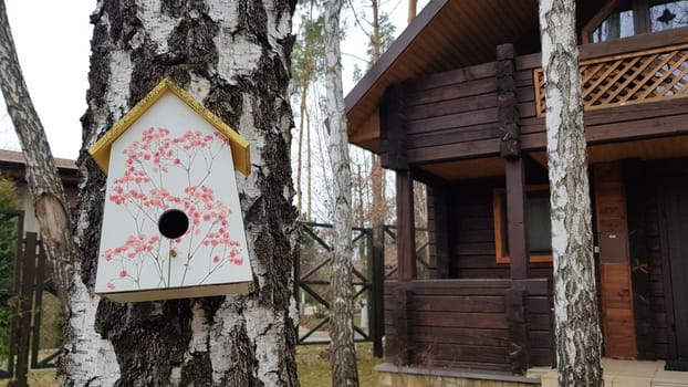 Ukraine, Kiev - April 17, 2020. Beautiful white painted birdhouse made of wood on a birch in the forest near a brown country wooden house. Homemade bird feeder. Art birdhouse on a tree in the garden