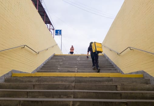 Ukraine, Kiev - September 26, 2019: A walking Glovo courier service provider with a yellow backpack rises the steps of the transition, hurries to deliver the order on time to the customer.