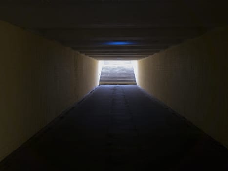 Empty yellow underground pedestrian crossing. Tunnel and daylight at the end. Steps to the top at the pedestrian crossing. A long concrete tunnel with lanterns in the city underground