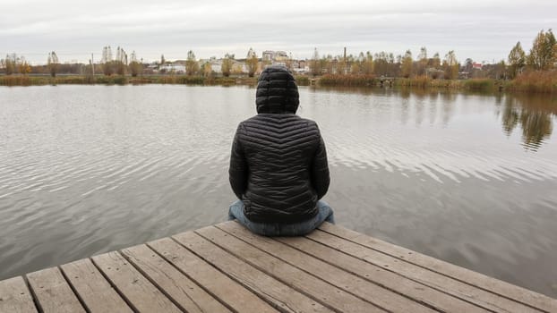 A girl in a jacket sits on a wooden bridge by the lake in cold autumn weather. Dipresia or dreaming alone. Thinking one on one with nature