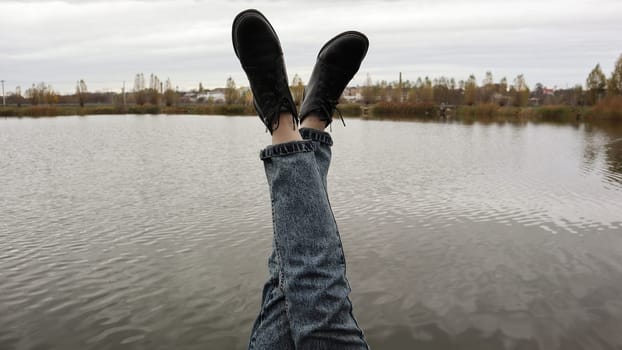 A girl in blue jeans sits by the lake. A young woman is resting by the river, sitting on the edge of a wooden bridge. Pair of legs relaxing by a lake on a wooden dock. People are resting