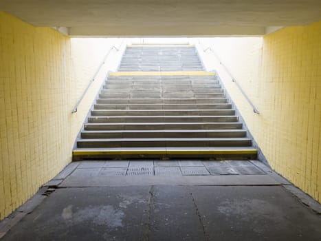 Empty yellow underground pedestrian crossing. Tunnel and daylight at the end. Steps to the top at the pedestrian crossing. A long concrete tunnel with lanterns in the city underground