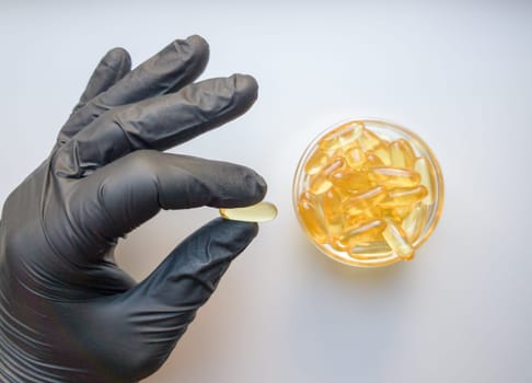 fish oil capsules in hand in a black medical glove on a white background, the hand takes one tablet from a saucer or plate. Hand holding omega 3 capsule on a white background. Healthcare concept.
