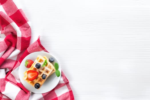 White plate on red kitchen towel with stack homemade Belgian waffles, blueberry, strawberry cuts, mint leaf, sugar powder on gray background