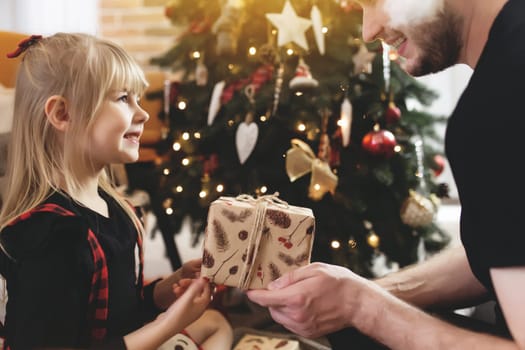 Young father in red Santa hat giving Christmas gift to his smiling daughter near decorated Christmas tree. Girl dressed in festive red-black Christmas outfit. They smiling, happy because of New Year