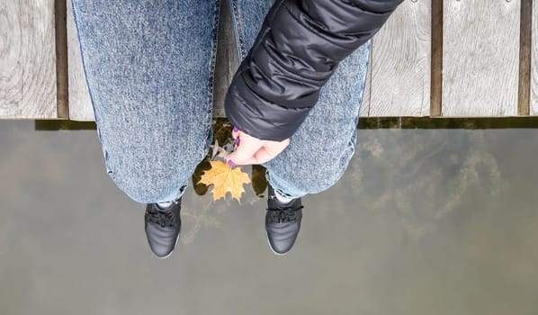 Beautiful legs of a girl in jeans are sitting on a wooden bridge by the lake, holding a yellow maple leaf in their hands. Autumn warm and sunny day. View from above. Autumn concept.