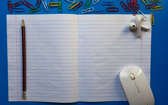 Flat lay photo of a school desk with a mouse, blank open notebook with pencil, paper clips, earphones on a blue wooden tabletop.