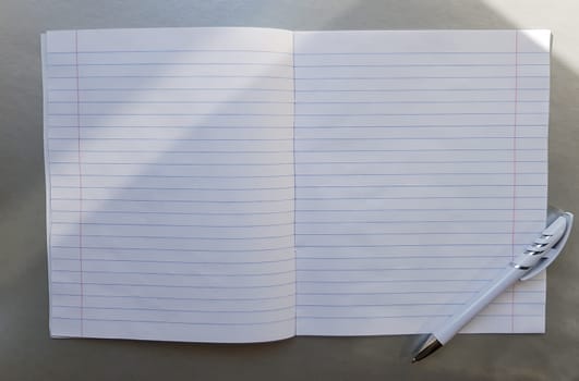 open school notebook with pen on a gray desk with sun rays from the window. A blank, white sheet of notebook for writing. education concept. Copy space. view from above. Flat lay.