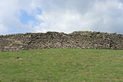 Plain countryside background image consisting of a green grassy field, long dry stone wall running through the middle and a clouded blue sky behind.