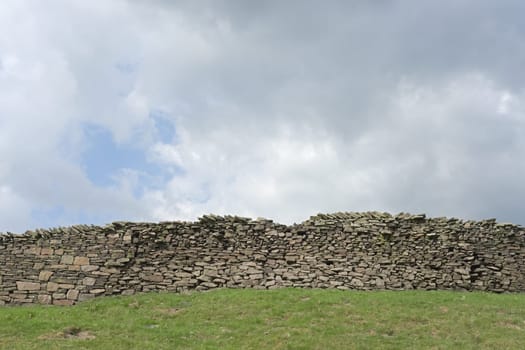 Large dry stone wall and a heavily clouded blue sky background. Agricultural and rural theme, green grassy field foreground. Plenty of copy space.