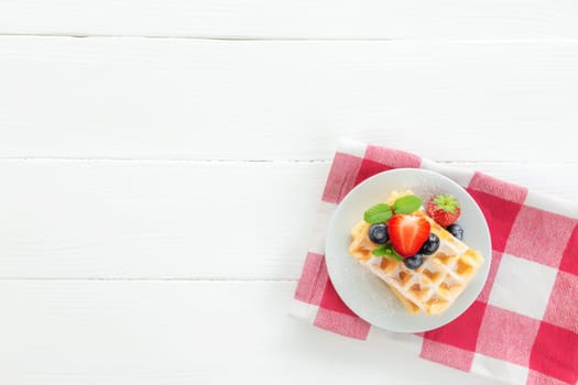 White plate on red kitchen towel with stack homemade Belgian waffles, blueberry, strawberry cuts, mint leaf, sugar powder on gray background