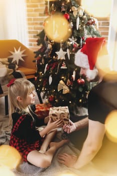 Young father in red Santa hat giving Christmas gift to his smiling daughter near decorated Christmas tree. Girl dressed in festive red-black Christmas outfit. They smiling, happy because of New Year