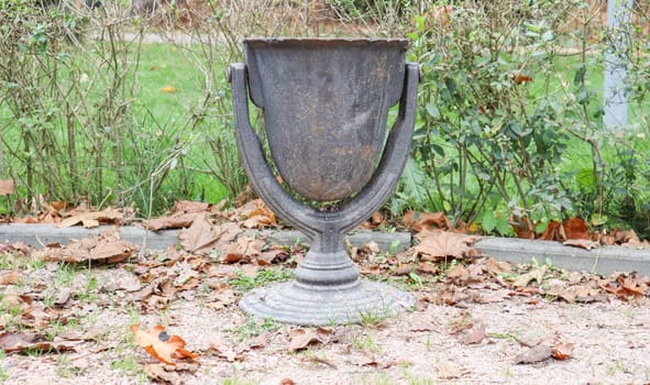 Old black curly garbage basket in a park in autumn. Public trash in a park surrounded by autumn leaves.