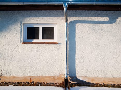 Detailansicht einer flachen Hausfassade mit Dachrinne, Fallrohr und kleines Fenster im seitlichen Abendlicht mit Schattenbild des Fallrohrs.