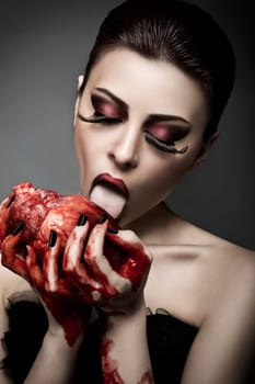 Beauty young woman licks blood from human heart against grey background