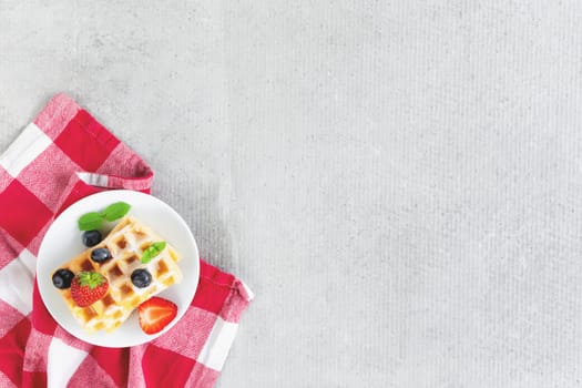 White plate on red kitchen towel with stack homemade Belgian waffles, blueberry, strawberry cuts, mint leaf, sugar powder on gray background