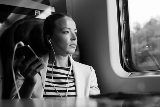 Thoughtful businesswoman looking trough the window, listening to podcast on cellphone using headphone set while traveling by train in business class seat. Black and white image.