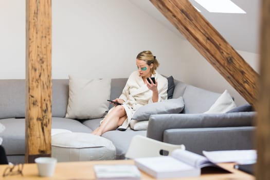 Young female entrepreneur wearing beauty face mask and cosy warm bathrobe in the morning, working remotly from her living room sofa. Remote work from home concept