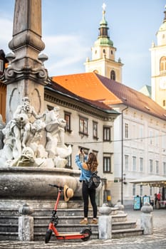 Traveler exploring old town on environmentally friendly electric scooter. Female tourist exploring Ljubljana's old medieval historical city center, taking picture of a fountain with her phone