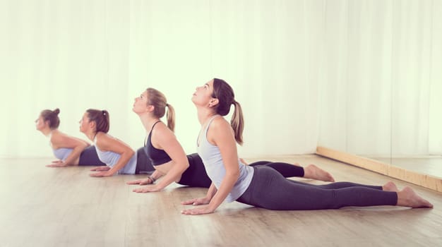 Group of young sporty sexy women in yoga studio, practicing yoga lesson with instructor, forming a line in asana pose. Healthy active lifestyle, working out indoors in gym.