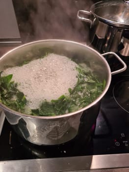 Spinach cooking preparation. Stewed spinach in black cooking pot with spoon on rustic background with ingredients and kitchen utensils . Top view. Home cuisine. Healthy food nutrition