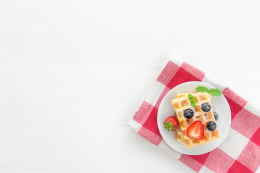 White plate on red kitchen towel with stack homemade Belgian waffles, blueberry, strawberry cuts, mint leaf, sugar powder on gray background