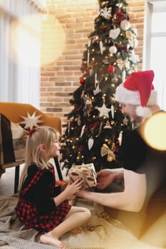 Young father in red Santa hat giving Christmas gift to his smiling daughter near decorated Christmas tree. Girl dressed in festive red-black Christmas outfit. They smiling, happy because of New Year