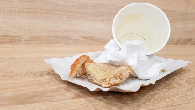 Leftover food on a paper plate and empty dirty paper coffee cup on a wooden table in a fast food cafe.