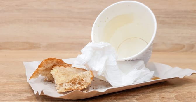 Leftover food on a paper plate and empty dirty paper coffee cup on a wooden table in a fast food cafe.