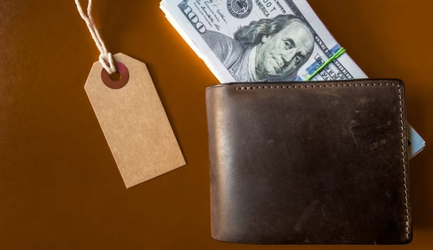 Many hundreds of dollars banknotes in a leather wallet with a brown tag, price tag on a brown background. Flat lay. Background, holiday concept. Black Friday - International Day of Shopping, Promotions, Discounts, Sales. Sales season is the fourth Thursday of November.