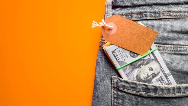 Many hundreds of dollars banknotes in a jeans pocket in a purse with a cardboard tag, price tag. Wallet and dollars in jeans pocket. Copy space, place for text, flat lay. Background, holiday concept. Black Friday - International Day of Shopping, Promotions, Discounts, Sales. Sales season is the fourth Thursday of November. Blue jeans on a colored background.