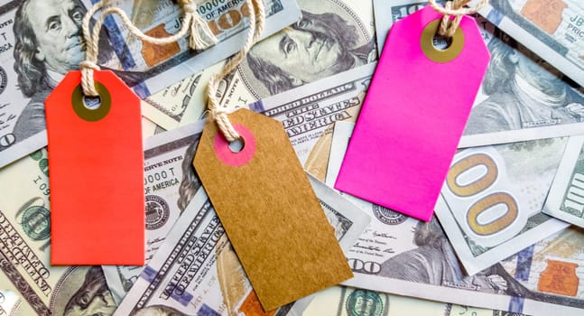 Three empty multi-colored and bright cardboard tags for the inscription Black Friday or discounts on the background of a heap of American hundred-dollar bills. Background, holiday concept. Black Friday - International day of purchases, promotions, discounts, sales. The sales season is the fourth Thursday of November. Copy space, place for text, flat lay.
