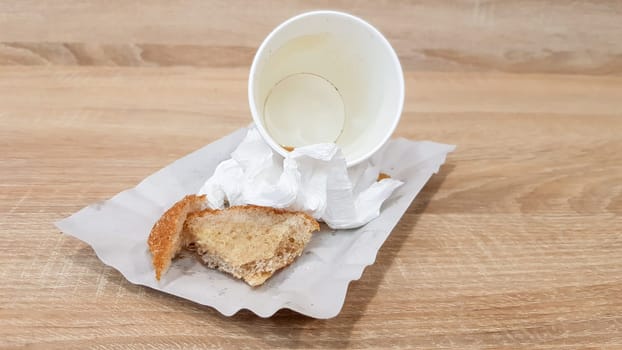 Leftover food on a paper plate and empty dirty paper coffee cup on a wooden table in a fast food cafe.