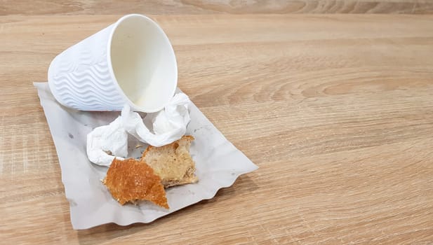 Leftover food on a paper plate and empty dirty paper coffee cup on a wooden table in a fast food cafe.