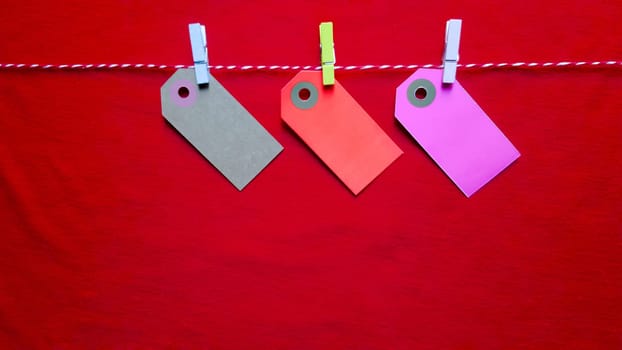 Colorful paper notes and clothespins on a red background. Empty color cardboard price tags, sale tag, gift tag, address label, luggage label hanging on clothes. Wooden clips. Layout, copy space for text, top view.