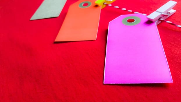 Colorful paper notes and clothespins on a red background. Empty color cardboard price tags, sale tag, gift tag, address label, luggage label hanging on clothes. Wooden clips. Layout, copy space for text, top view.