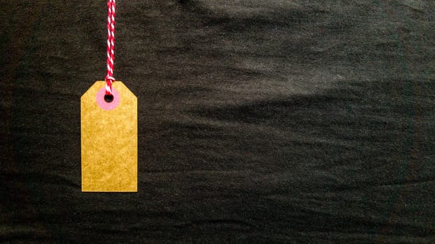 Brown cardboard tag hanging on a red rope on a black background from fabric. Copy space, place for text, flat lay