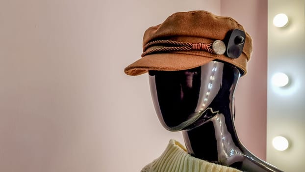 Portrait of a black mannequin with a brown hat in a women fashion store. Mannequin woman in a hat on a pink wall background and a large white mirror with backlight and light bulbs.