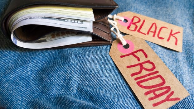 Brown wallet with money, dollars on a blue background, jeans texture, cardboard tag with red text Black Friday. Black Friday - International day of purchases, promotions, discounts, sales. Copy space, place for text, flat lay.