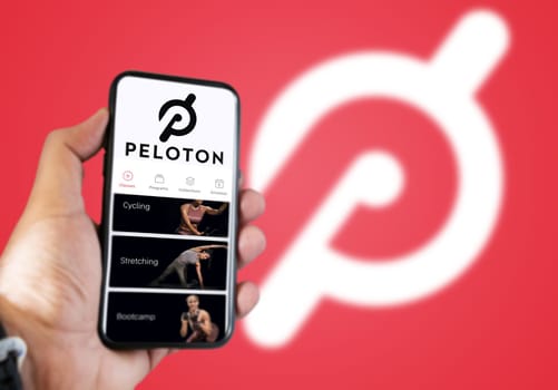 New York, USA, July 2021: A hand holding a phone with the Peloton app on the screen. Red background with blurred Peloton logo. Peloton is an American exercise equipment based in New York