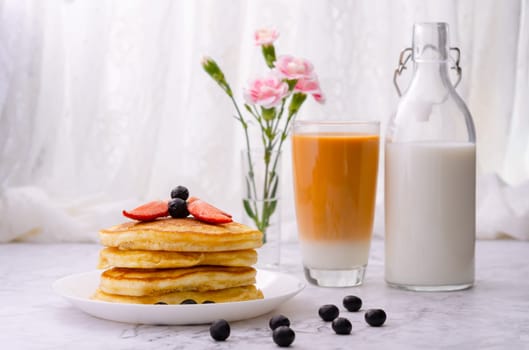 Stack of pancakes with fresh strawberries and blueberries on a white plate, a bottle of milk, a glass of milk tea and a vase of pink flowers on marble table. Healthy morning breakfast.