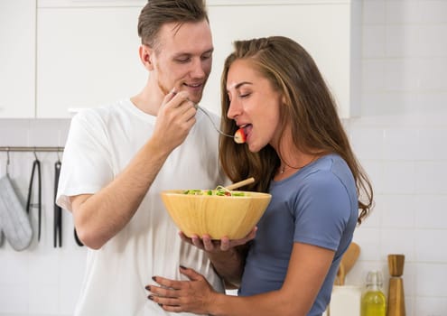 Happy young couple enjoying and preparing a healthy meal in home kitchen. Man holding salad bowl and offering a strawberry to woman