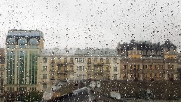 Wet window with drops on the background of the autumn city in cloudy weather. View from the window in the rain. Water drop on glass window during rain with blurred background of city scene.