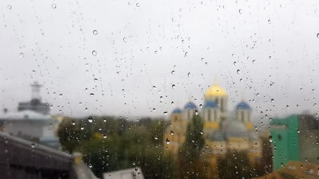Wet window with drops on the background of the autumn city in cloudy weather. View from the window in the rain. Water drop on glass window during rain with blurred background of city scene.