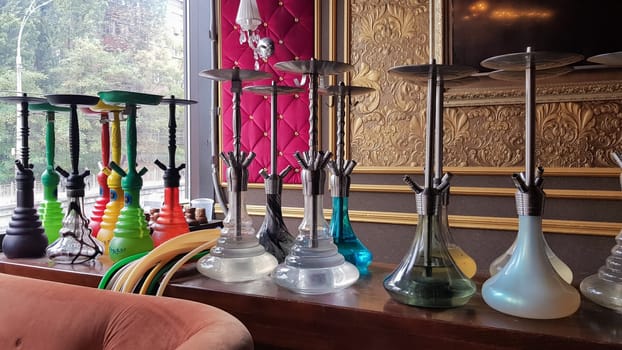 Group of modern glass hookah appliance on a wooden stand. Restaurant bar. Glass hookahs on the bar in a cafe or bar made of beautiful multi-colored glass. Diffused daylight from the window