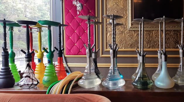 Group of modern glass hookah appliance on a wooden stand. Restaurant bar. Glass hookahs on the bar in a cafe or bar made of beautiful multi-colored glass. Diffused daylight from the window