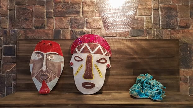 Ukraine, Kiev - June 10, 2020. Ceramic figurines and figures decoration or symbol in the home interior. Two masks on a wooden shelf against the background of a stone wall with backlight.