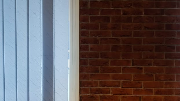 Large window with vertical white or gray blinds, loft, brick wall, industrial interior. Sunlight from a window on brick walls in a room or office