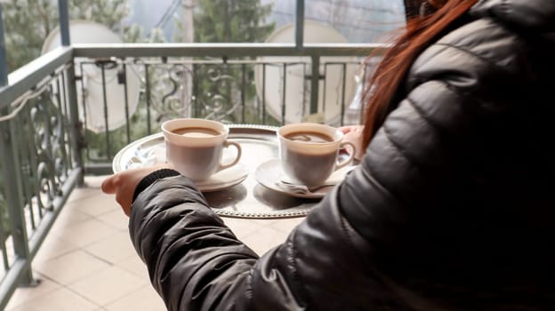 two cups of coffee on a tray in the hands of a girl. A woman with cups in her hands enters the hotel's balcony in the morning. Drink coffee with mountain and forest views