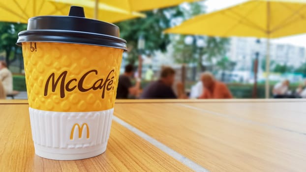 McCafe menu at McDonalds restaurant. Yellow cup of coffee on a table outdoors with yellow umbrellas and visitors in the background. Breakfast, coffee time. Ukraine, Kiev - September 6, 2019